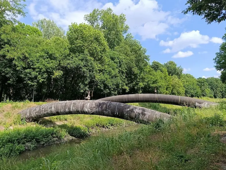 A large pipeline crosses a small stream in a green, wooded landscape under a partly cloudy sky. The image illustrates the intersection of infrastructure, the natural environment and the challenges of sustainable development.