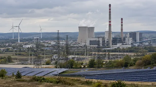 Industrial energy landscape with a conventional power plant, electricity transmission lines, wind turbines and solar panels under an overcast sky, illustrating the coexistence of fossil fuels and renewable energy.